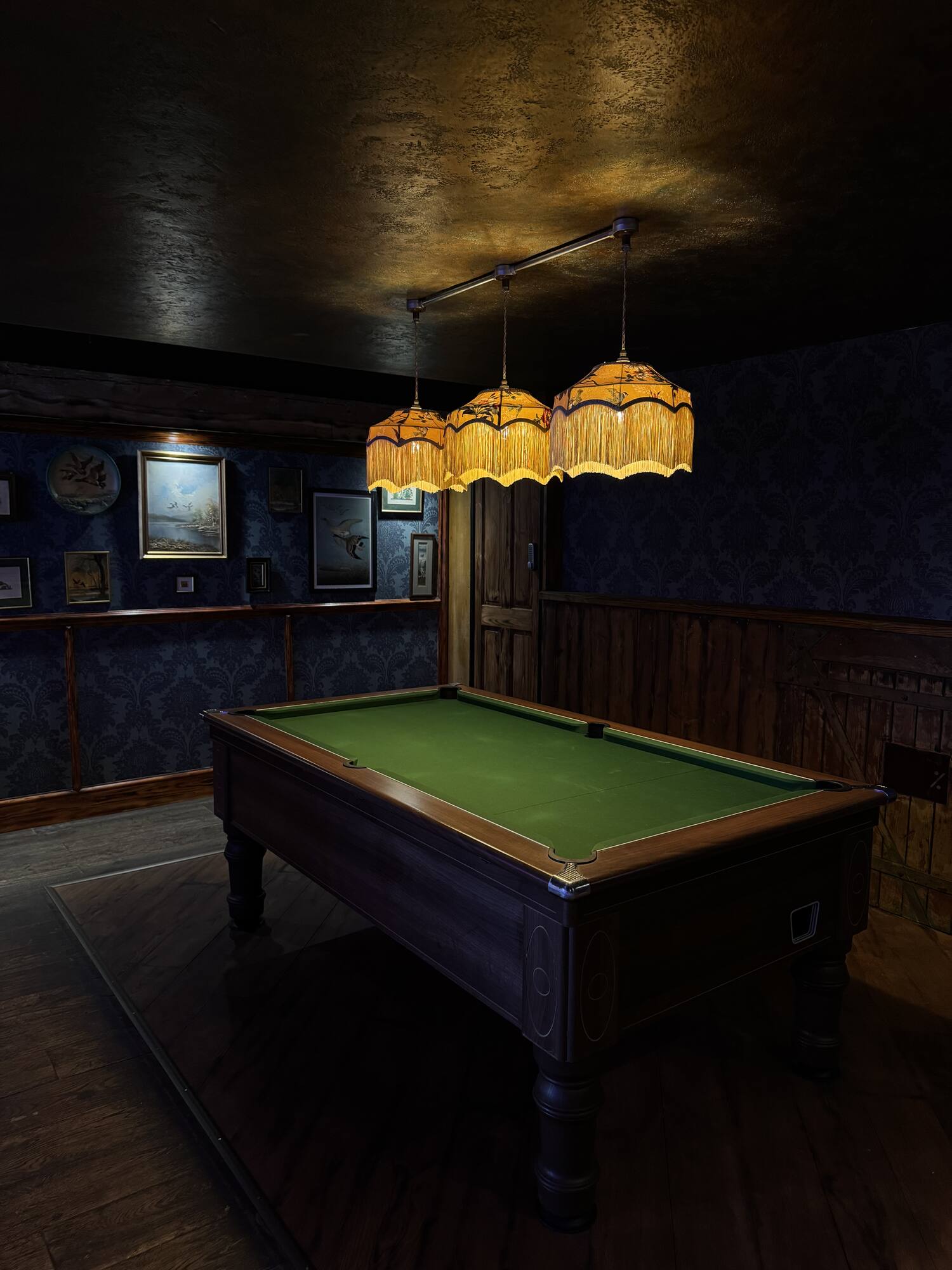 A pool table with green baize beneath fringed pendant lamps and framed prints