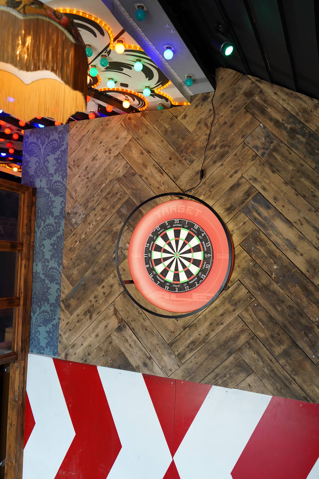 The interactive dartboard glowing red on a herringbone wood wall above a chevron-painted oche