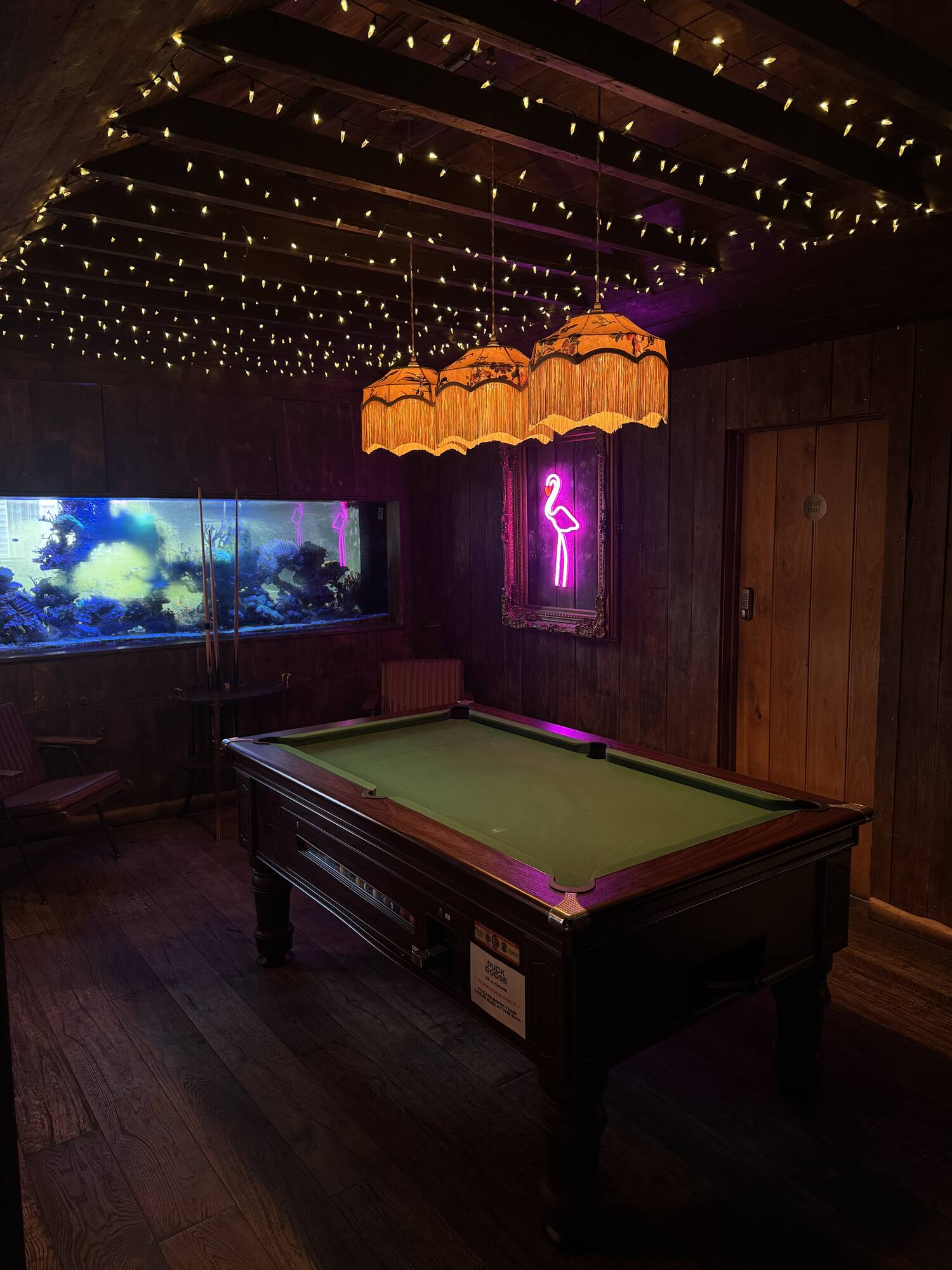A pool table by a lit fish tank and neon flamingo, strung with fairy lights